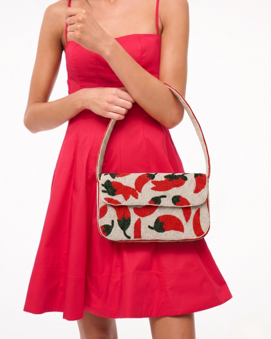 Model in a coral-red dress carries The Cayenne, a cream hand-beaded shoulder bag scattered with red chili peppers and green leaves.