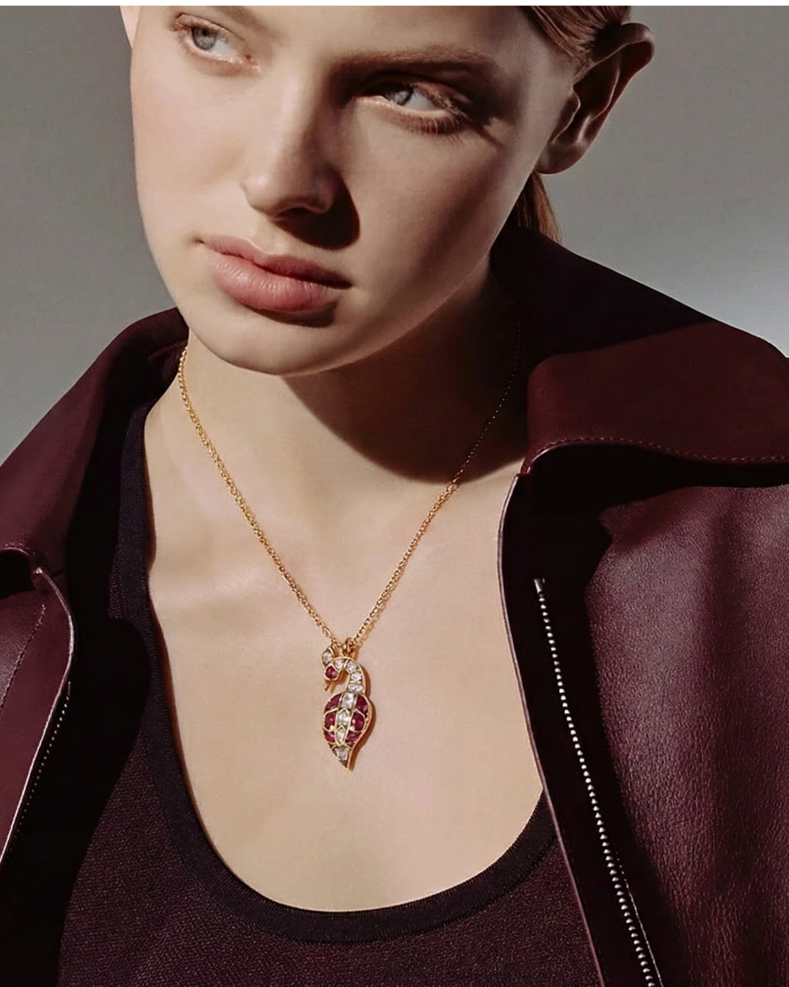 Model in a burgundy jacket wears The Crimson Swan Pendant, a gold paisley pendant set with ruby and crystal stones.