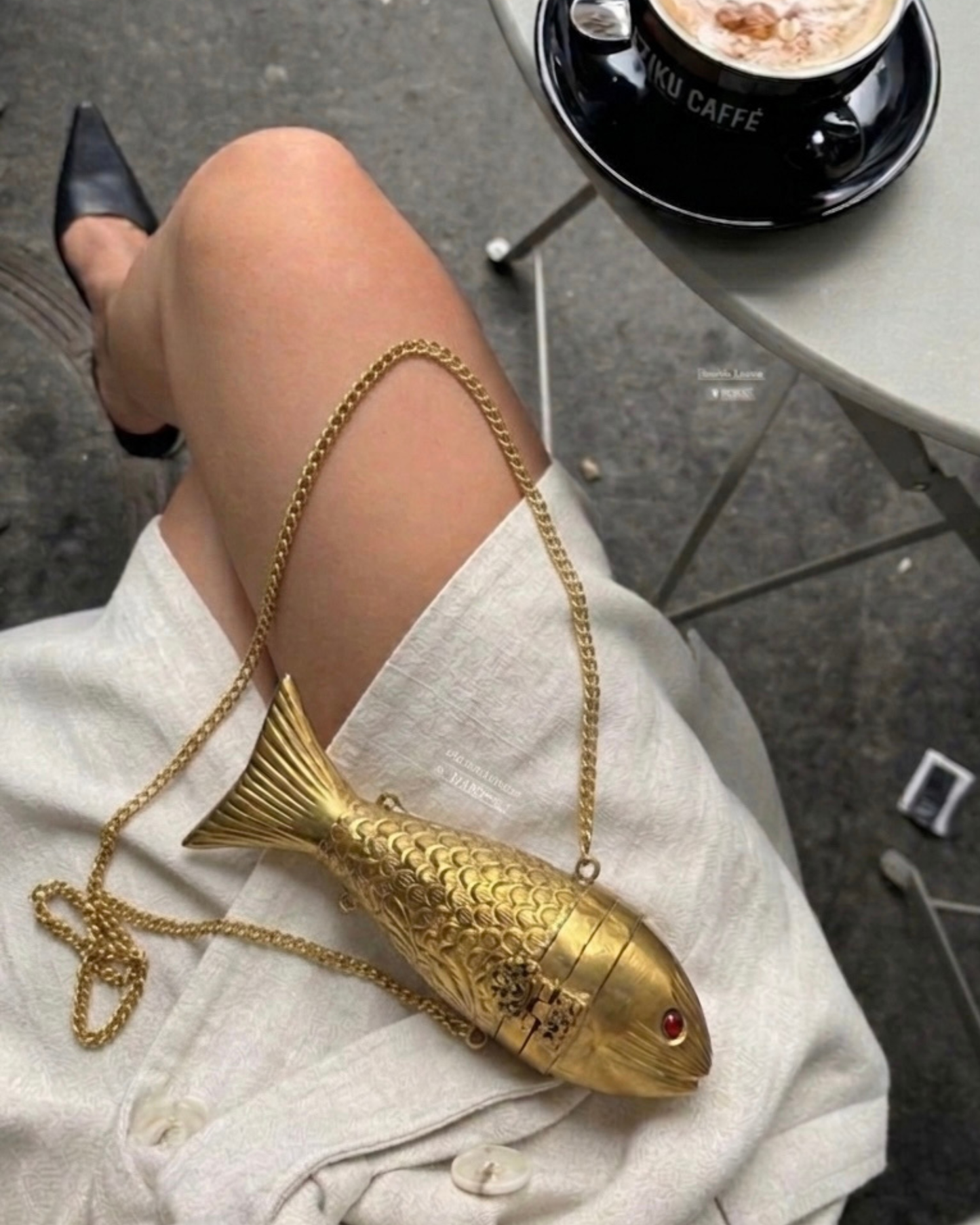 The Siren gold fish minaudière resting on a lap with its chain strap, at a cafe table.