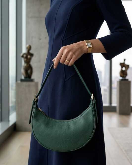 Model in a navy dress holds The Ivy, a forest-green crescent shoulder bag in smooth leather.